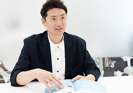 赤い水玉ネクタイを着けた男性社員がパンフレットを広げて笑顔で対応する様子